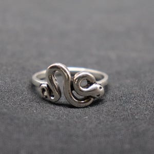 May include: A silver-toned metal ring featuring a coiled snake design. The snake body forms the band of the ring, with the head and tail meeting at the front. The metal has a polished finish with darkened recessed areas to highlight the snake shape.