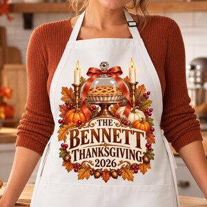 May include: A white apron featuring a graphic of a pie under a glass dome, flanked by two candles, pumpkins, and autumn leaves. The text reads THE BENNETT THANKSGIVING 2026, surrounded by a decorative border of fall foliage, berries, and acorns.