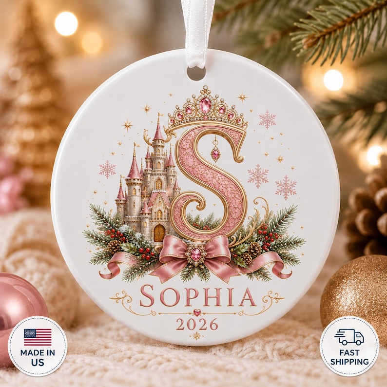 Op de afbeelding: Een rond wit keramieken ornament met een grote roze letter S bekroond met een juwelenkroon. Een sprookjeskasteel, dennentakken, rode bessen en een roze lint zijn eronder afgebeeld. De naam Sophia en het jaartal 2026 zijn in het roze gedrukt.