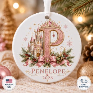 Op de afbeelding: Een ronde witte keramische hanger met een grote roze letter P versierd met een tiara, een gedetailleerd kasteel, dennentakken, rode bessen en een roze strik. De tekst luidt Penelope en 2026. Een wit lint is aan de bovenkant bevestigd.