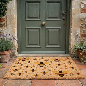 May include: A rectangular coir doormat with a repeating pattern of bees and flowers in yellow, orange, and black. The mat is placed in front of a green door. Potted plants are visible on either side of the door.
