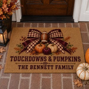 May include: A rectangular tan coir doormat featuring a large maroon and tan plaid bow centered above a football, three pumpkins, and autumn foliage. The text reads TOUCHDOWNS & PUMPKINS and THE BENNETT FAMILY in dark maroon block lettering.