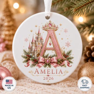 Op de afbeelding: Een rond wit keramieken ornament met een grote roze letter A bekroond met een juwelenkroon. Naast de letter staat een gedetailleerd kasteel, omringd door dennentakken, rode bessen en een roze strik. De naam AMELIA en het jaartal 2026 staan eronder.