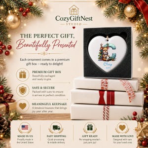 Op de afbeelding: Een wit hartvormig keramisch ornament met een illustratie van een rendier, de letter C en de naam Cindy 2025. Het ornament wordt gepresenteerd in een zwarte geschenkdoos met schuimvoering op een stapel van drie witte geschenkdozen.