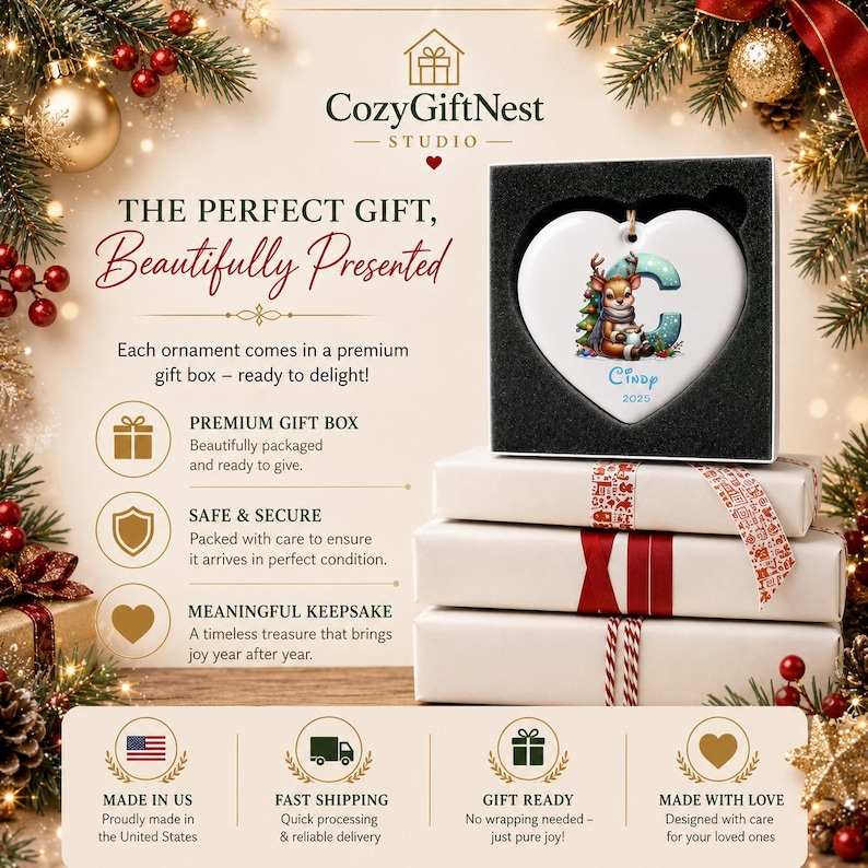 Op de afbeelding: Een wit hartvormig keramisch ornament met een illustratie van een rendier, de letter C en de naam Cindy 2025. Het ornament wordt gepresenteerd in een zwarte geschenkdoos met schuimvoering op een stapel van drie witte geschenkdozen.