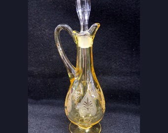 May include: Yellow glass cruet with a handle and a clear stopper. The cruet has etched floral designs and a round base. The stopper is clear and pointed. The cruet is approximately 8 inches tall.