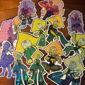 Puede incluir: Una colección de pegatinas coloridas con personajes de dibujos animados en varias poses. Las pegatinas muestran personajes con tonos de piel amarillos, verdes, morados y naranjas. Algunas pegatinas tienen un efecto holográfico, añadiendo brillo a los diseños.