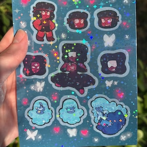 Peut inclure: Une planche d'autocollants holographiques représentant des personnages de dessins animés rouges, noirs et bleus. Les autocollants sont sur un fond bleu sarcelle avec des cœurs, des étoiles et des papillons. Les autocollants sont de tailles et de formes variées.