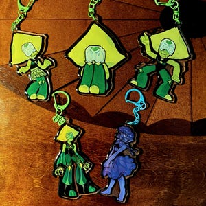 Puede incluir: Llaveros con personajes de dibujos animados en tonos verdes y azules. Los llaveros tienen un cierre verde y un diseño de personaje de dibujos animados. Los personajes están en varias poses, con una combinación de colores amarillo-verde.