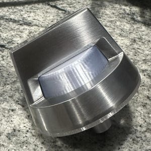 Può includere: Manopola per elettrodomestici in metallo spazzolato con un design arrotondato e angolato. La manopola ha un inserto blu chiaro, semitrasparente e una base cilindrica. La superficie presenta segni di lavorazione visibili, conferendole un aspetto strutturato.