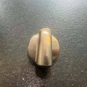 Könnte beinhalten: Ein Knopf aus gebürstetem Metall mit zylindrischem Griff und kreisförmiger Basis. Der Knopf hat eine hellsilberne Farbe und scheint aus Edelstahl zu bestehen. Der Hintergrund ist eine dunkle, strukturierte Oberfläche.