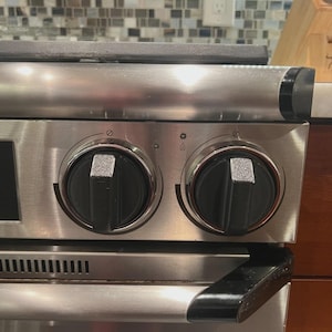 May include: Close-up of a stainless steel gas range with two black control knobs. The knobs have a silver accent and are labeled with symbols. The range has a black handle and a vent along the bottom.