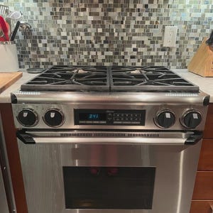 May include: Stainless steel gas range with four burners and an oven. The range has black control knobs and a digital display showing the time. The oven door has a handle and a glass window. The backsplash is tiled with small, square tiles.
