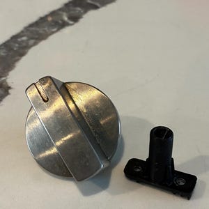 May include: A stainless steel oven knob with a ridged grip and a separate black plastic base. The knob is circular with a flat top and a curved, ridged handle. The black base has a rectangular shape with two holes.