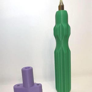 May include: A green screwdriver with a gold tip stands upright next to a purple T-shaped object with two holes. The screwdriver has a ribbed handle. The objects are on a white surface.