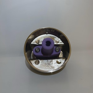 May include: A circular brass-coloured component featuring a central purple plastic piece. The purple part has a cylindrical hole and is secured with screws. This appears to be a mechanical part or fitting.