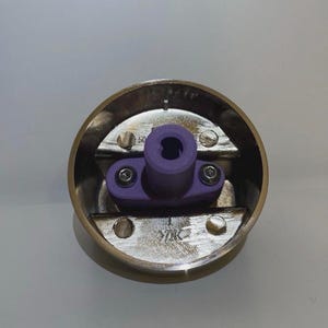 Könnte beinhalten: Nahaufnahme eines kreisförmigen, messingfarbenen Objekts mit einem zentralen lila Kunststoffteil, das mit Schrauben befestigt ist. Das Objekt hat eine metallische Oberfläche und scheint ein mechanisches Teil zu sein.
