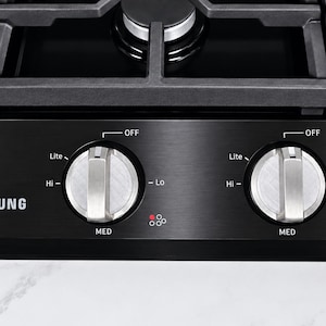 May include: A black Samsung gas cooktop control panel featuring two silver metallic knobs with settings for Lite, Hi, Med, Lo, and Off. The panel has a brushed metal finish with white text and red indicator dots for burner positions.