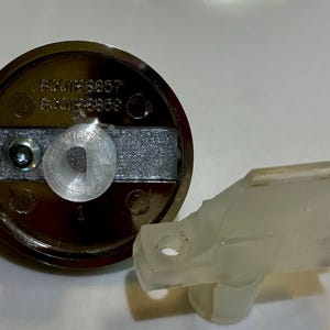 May include: A circular dark brown control knob featuring a central silver-toned metal bar with a small screw and a translucent plastic insert. Beside it lies a separate, L-shaped translucent plastic component with a small circular opening.