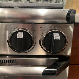 Può includere: Primo piano di un fornello a gas in acciaio inossidabile con due manopole di controllo nere. Le manopole hanno un accento argentato e sono posizionate su una superficie in metallo spazzolato. Il fornello presenta una maniglia nera e una ventilazione.