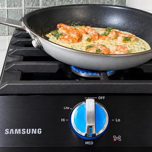 May include: A black Samsung gas cooktop with a blue control knob featuring settings for Lite, Hi, Med, and Lo. A silver frying pan with a blue handle sits on the black metal grate, containing cooked shrimp in a creamy sauce with green herbs.