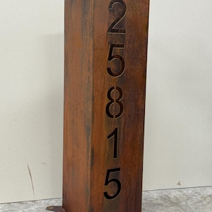 May include: A rectangular, weathered metal address sign with the numbers 25815 cut out. The sign has a rusty brown color and a square base with mounting holes. The numbers are vertically aligned.