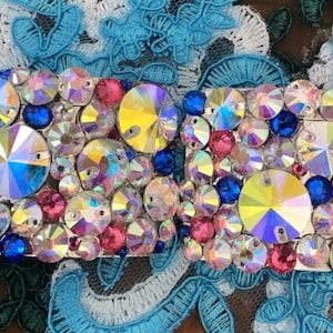 May include: A bedazzled accessory featuring an array of colorful rhinestones in shades of blue, pink, and yellow. The sparkling embellishments are set against a white base, creating a glamorous and eye-catching design. The background includes blue and white lace.