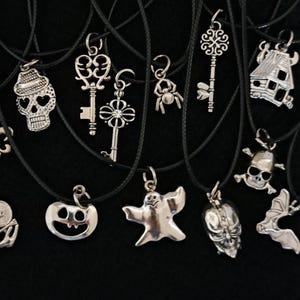 May include: A collection of silver-toned pendant necklaces with various charms, including keys, a skull, a spider, a ghost, and a pumpkin. The pendants hang on black cords against a dark background.