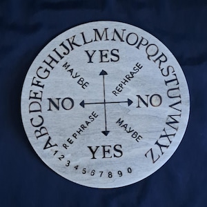 Puede incluir: Un tablero de Ouija redondo de madera clara con letras, números e indicadores direccionales grabados. El tablero presenta las palabras "SÍ", "NO", "QUIZÁS" y "REFORMULAR", junto con el alfabeto y los números 1-9 y 0.