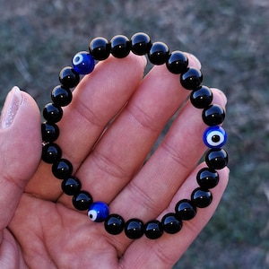 May include: Black onyx bead bracelet with three blue and white evil eye beads. The bracelet is made of polished, round black beads strung together with elastic cord. The evil eye beads are a vibrant blue with a white iris and black pupil.