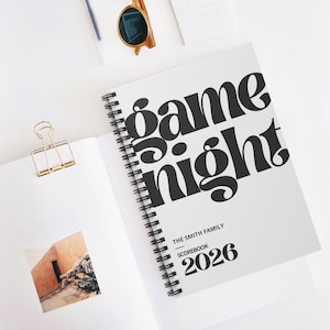 Puede incluir: Un cuaderno blanco con espiral con las palabras "game night" en letras negras grandes. El texto "THE SMITH FAMILY SCOREBOOK 2026" está impreso debajo. Un clip de papel dorado y gafas de sol son visibles.