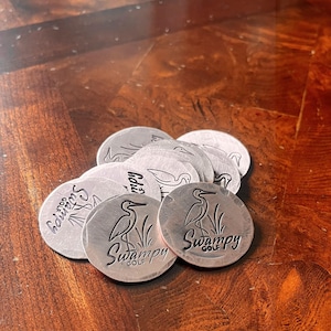 May include: A collection of silver-colored golf ball markers. Each marker features a heron illustration and the words "Swampy Golf" in a circular design. The markers are arranged on a dark wooden surface.