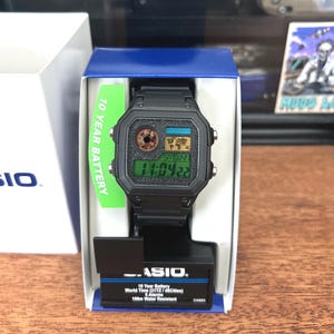 Puede incluir: Reloj digital negro con esfera cuadrada, presentado en su caja original. La esfera muestra la hora y la fecha, junto con un mapa del mundo. La caja incluye una etiqueta verde que dice "10 YEAR BATTERY".