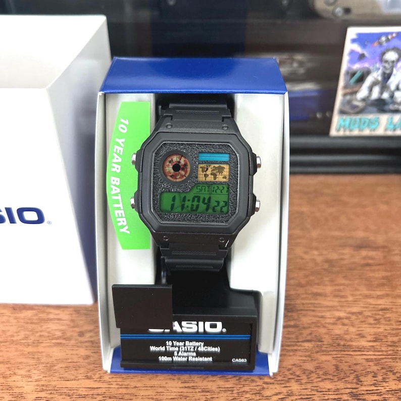 Puede incluir: Reloj digital negro con esfera cuadrada, presentado en su caja original. La esfera muestra la hora y la fecha, junto con un mapa del mundo. La caja incluye una etiqueta verde que dice "10 YEAR BATTERY".