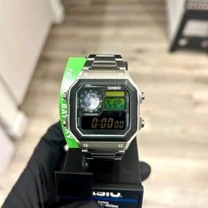 May include: A silver Casio digital watch with a black display showing the time as 0:00:00. The watch has a rectangular face with buttons and a metal band. The watch is on a black display stand.