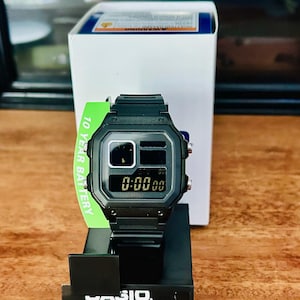 Könnte beinhalten: Schwarze Digitaluhr mit rechteckigem Zifferblatt, das "0:00:00" anzeigt. Die Uhr hat ein schwarzes Armband und steht auf einem schwarzen Präsentationsständer. Ein grünes Etikett zeigt "10 YEAR BATTERY". Die Uhr ist wasserdicht.