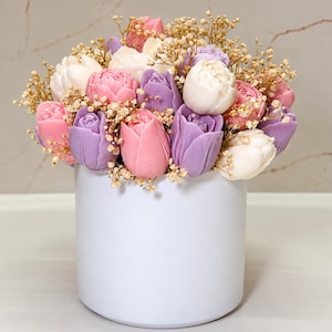 May include: A cylindrical white container filled with a dense arrangement of tulip-shaped flowers in shades of pastel pink, lavender, and white, interspersed with small sprigs of dried baby's breath filler flowers throughout the bouquet.