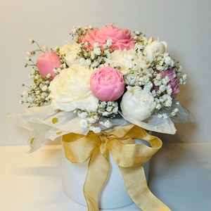 Puede incluir: Un arreglo floral con velas en forma de peon&iacute;as rosas y blancas, acentuado con delicado paniculata blanca. Las velas est&aacute;n dispuestas en una caja redonda blanca, envueltas en papel de seda y atadas con una cinta dorada.