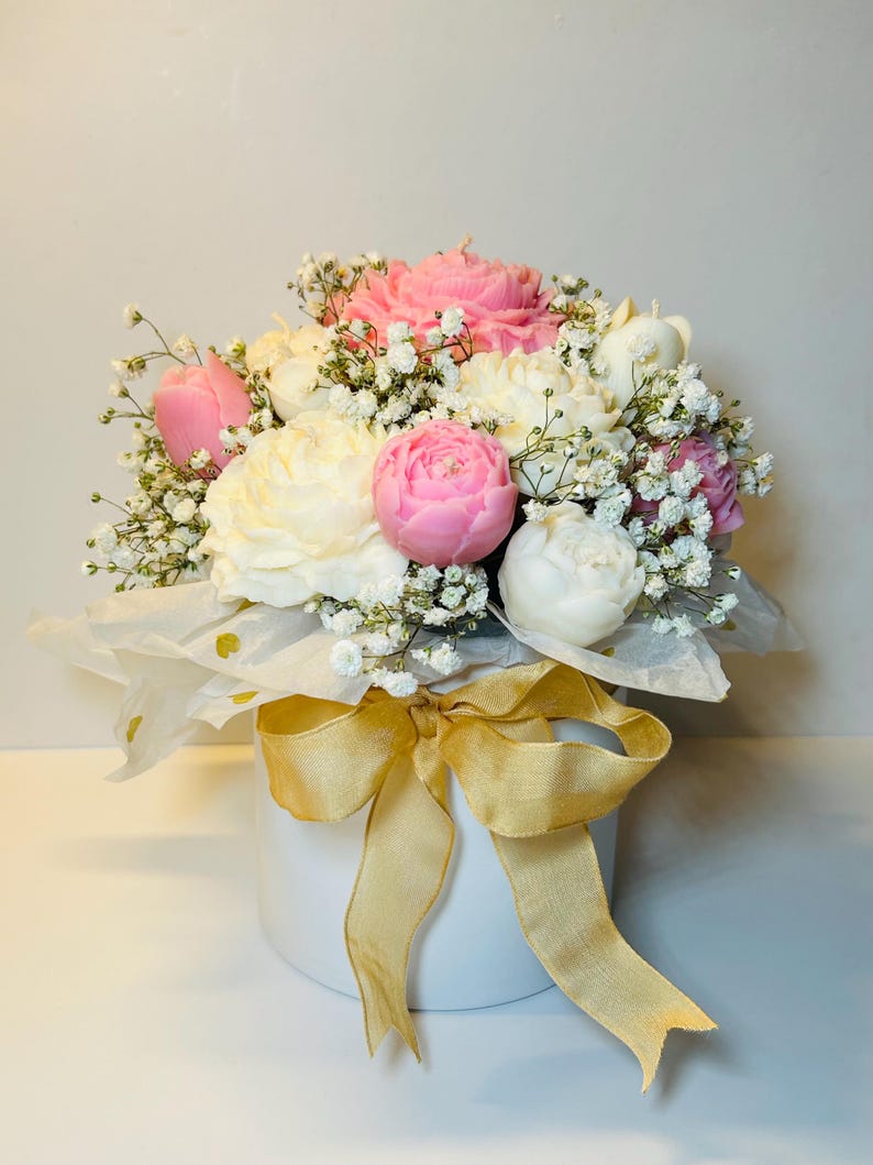 Puede incluir: Un arreglo floral con velas en forma de peon&iacute;as rosas y blancas, acentuado con delicado paniculata blanca. Las velas est&aacute;n dispuestas en una caja redonda blanca, envueltas en papel de seda y atadas con una cinta dorada.