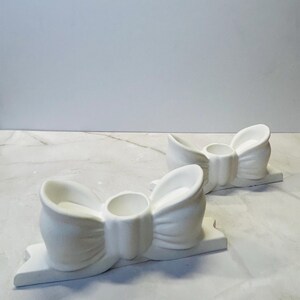 Può includere: Due portacandele bianchi in ceramica a forma di fiocco. Ogni supporto ha un'apertura circolare centrale per una candela e poggia su una base rettangolare. I fiocchi presentano pieghe e curve dettagliate.