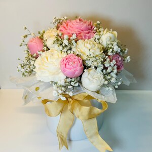 Puede incluir: Un ramo de peon&iacute;as rosas, blancas y crema y paniculata en un recipiente redondo blanco. El arreglo est&aacute; envuelto en papel de seda blanco y atado con una cinta dorada.
