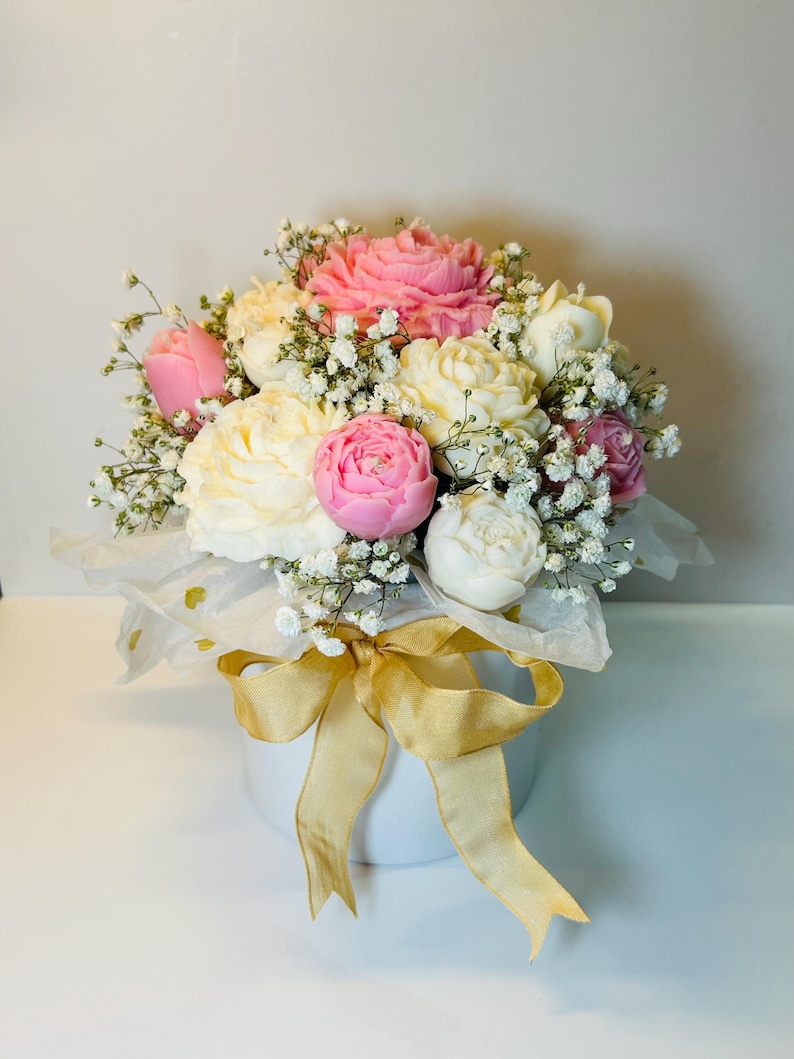 Puede incluir: Un ramo de peon&iacute;as rosas, blancas y crema y paniculata en un recipiente redondo blanco. El arreglo est&aacute; envuelto en papel de seda blanco y atado con una cinta dorada.