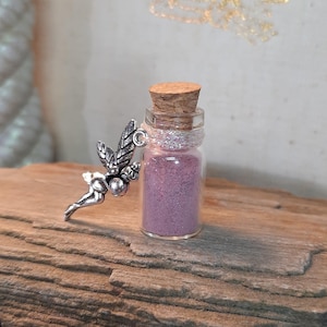 May include: A small glass bottle filled with purple sand, topped with a cork, and adorned with a silver fairy charm. The fairy has detailed wings and is attached to the bottle's neck. The bottle sits on a piece of wood.