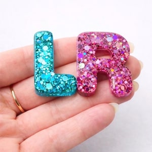 May include: Two glitter-filled resin letters, 'L' in teal and 'R' in pink, are displayed. The letters are filled with sparkling glitter, creating a shimmering effect. The letters are held in a hand, showcasing the vibrant colors and textures.