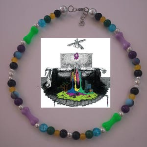 May include: A necklace with a central graphic and colourful beads. The necklace features a variety of beads in colours like green, blue, purple, yellow, and black, with bone-shaped green and purple accents. The graphic is a black and white illustration.