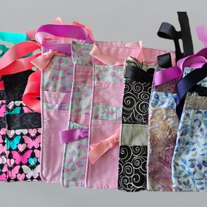 May include: A collection of colorful tote bags with various patterns, including butterflies, swirls, and floral designs. Each bag features a different color scheme, with ribbons and bows adding decorative accents. The bags are made of fabric and have handles.