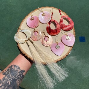 May include: A collection of assorted earrings displayed on a circular wood slice. The set includes pink and white shell-like circular and heart-shaped drops, some featuring small turtle or lotus charms, and two long earrings with cream-colored tassels.