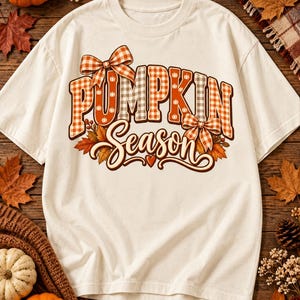 Peut inclure: Un t-shirt à manches courtes de couleur crème avec une impression graphique portant le texte "PUMPKIN SEASON" dans un motif vichy orange et marron. Le design comprend deux nœuds à carreaux, des icônes de citrouille et des feuilles d'automne.