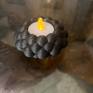 May include: A black, 3D-printed candle holder with a textured, braided design. The holder surrounds a white candle with a flickering, yellow LED light. The candle holder is sitting on a reflective surface.