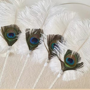 May include: Four white feather fans with peacock feather accents. Each fan features a large cluster of white feathers and a single peacock feather with blue, green, and brown hues. The handles are silver with rhinestone embellishments.